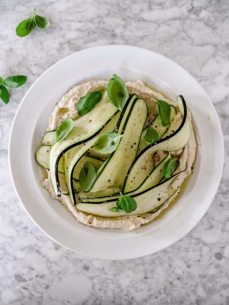 Hvid bønnecreme smurt ud på et lyst fad og toppet med tynde bånd af rå squash, frisk basilikum, citronskal, sort peber og olivenolie